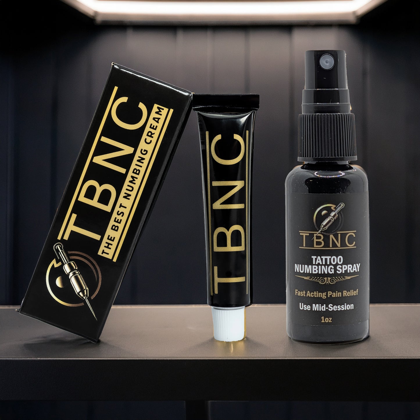 The TBNC - The Best Numbing Cream Tattoo Numbing Bundle features a black and gold numbing cream and a black numbing spray, both elegantly displayed on a dark surface against a sleek black background.