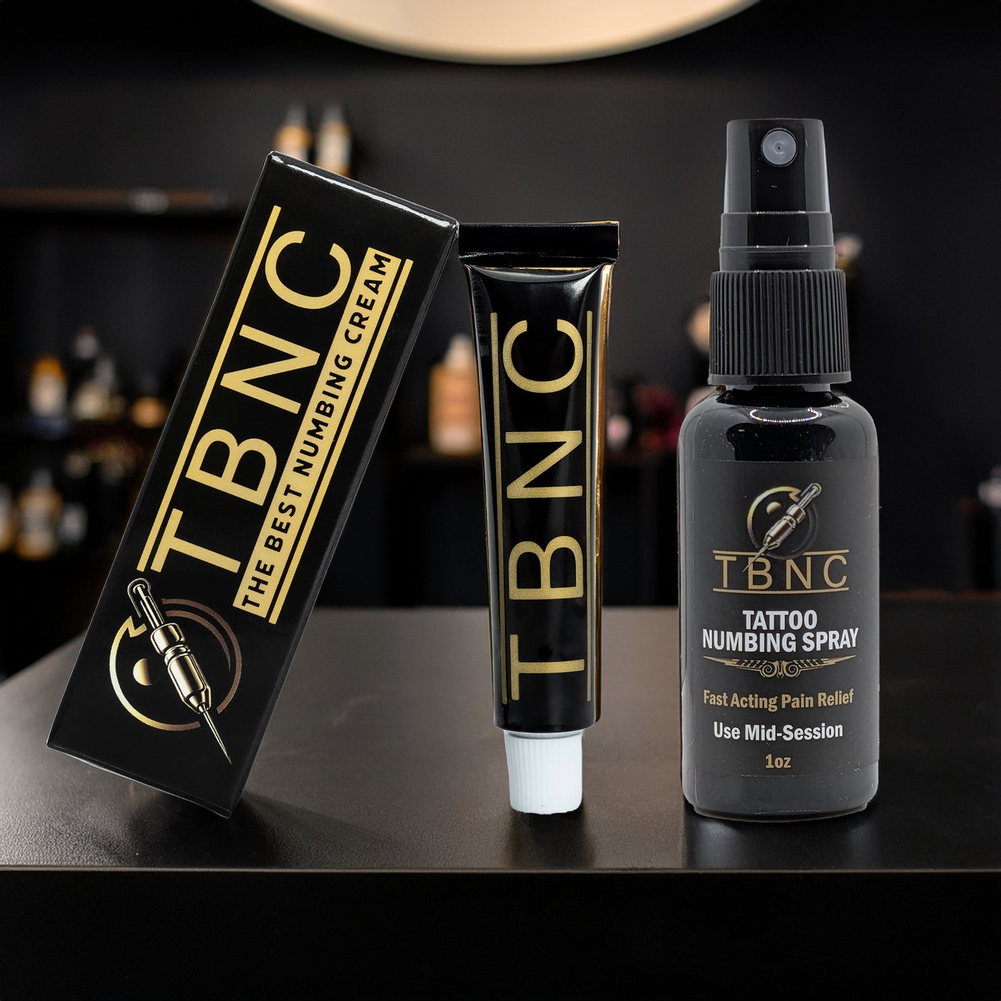 A black tube and box labeled "TBNC" from the Tattoo Numbing Bundle by TBNC - The Best Numbing Cream sit next to a black bottle marked "TBNC Tattoo Numbing Spray" on a dark surface with a blurred background.