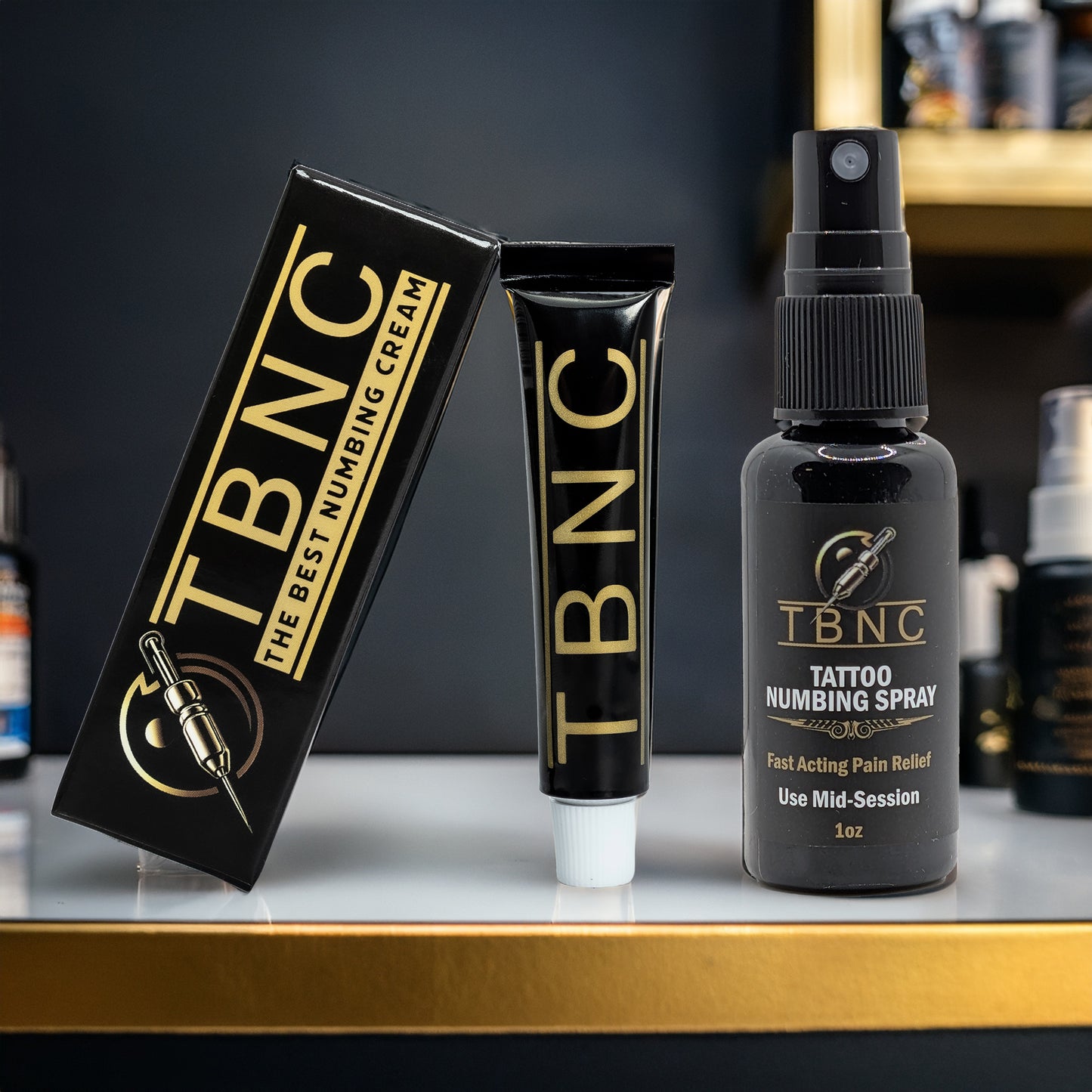 A tube and box labeled "TBNC - The Best Numbing Cream" sit beside a black Tattoo Numbing Spray bottle, both from the TBNC Tattoo Numbing Bundle, displayed on a shelf with blurred products in the background.