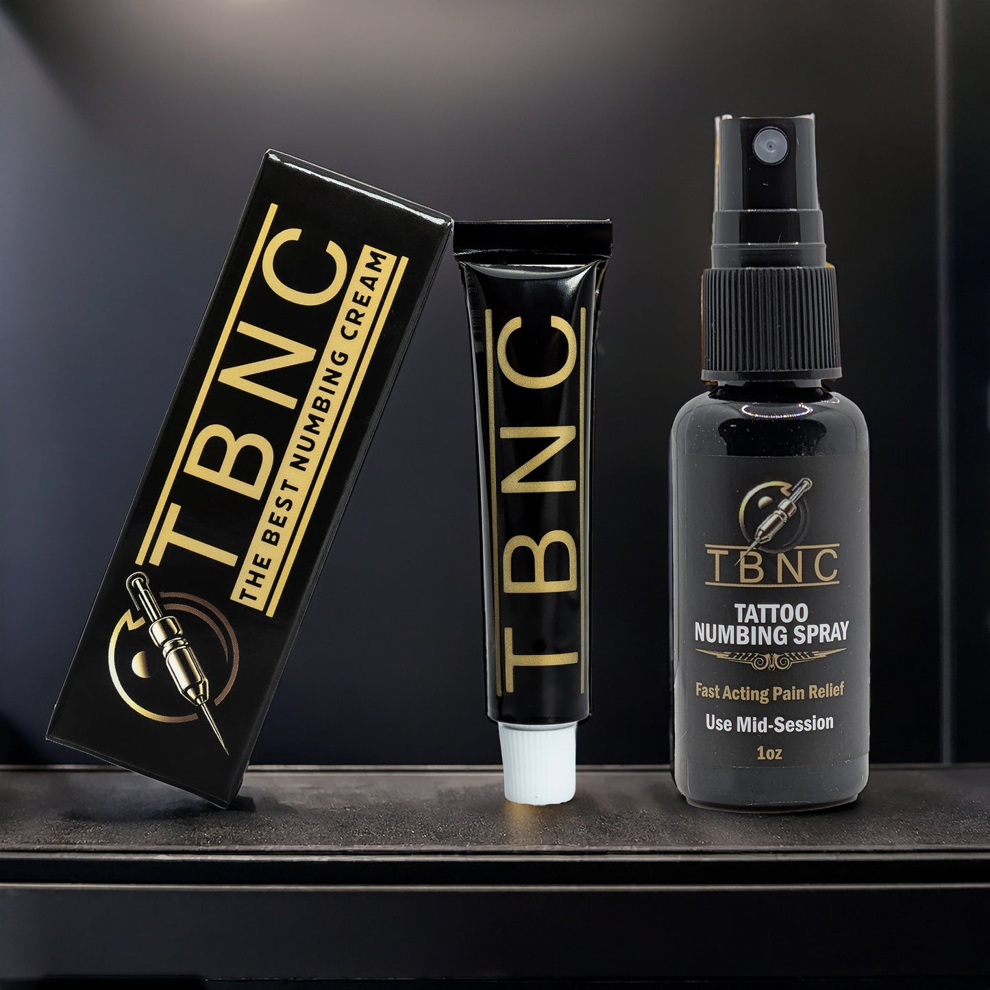 Three TBNC - The Best Numbing Cream products—Tattoo Numbing Cream in a black and gold box, a matching tube, and Tattoo Numbing Spray—are shown on a dark shelf as the Tattoo Numbing Bundle.