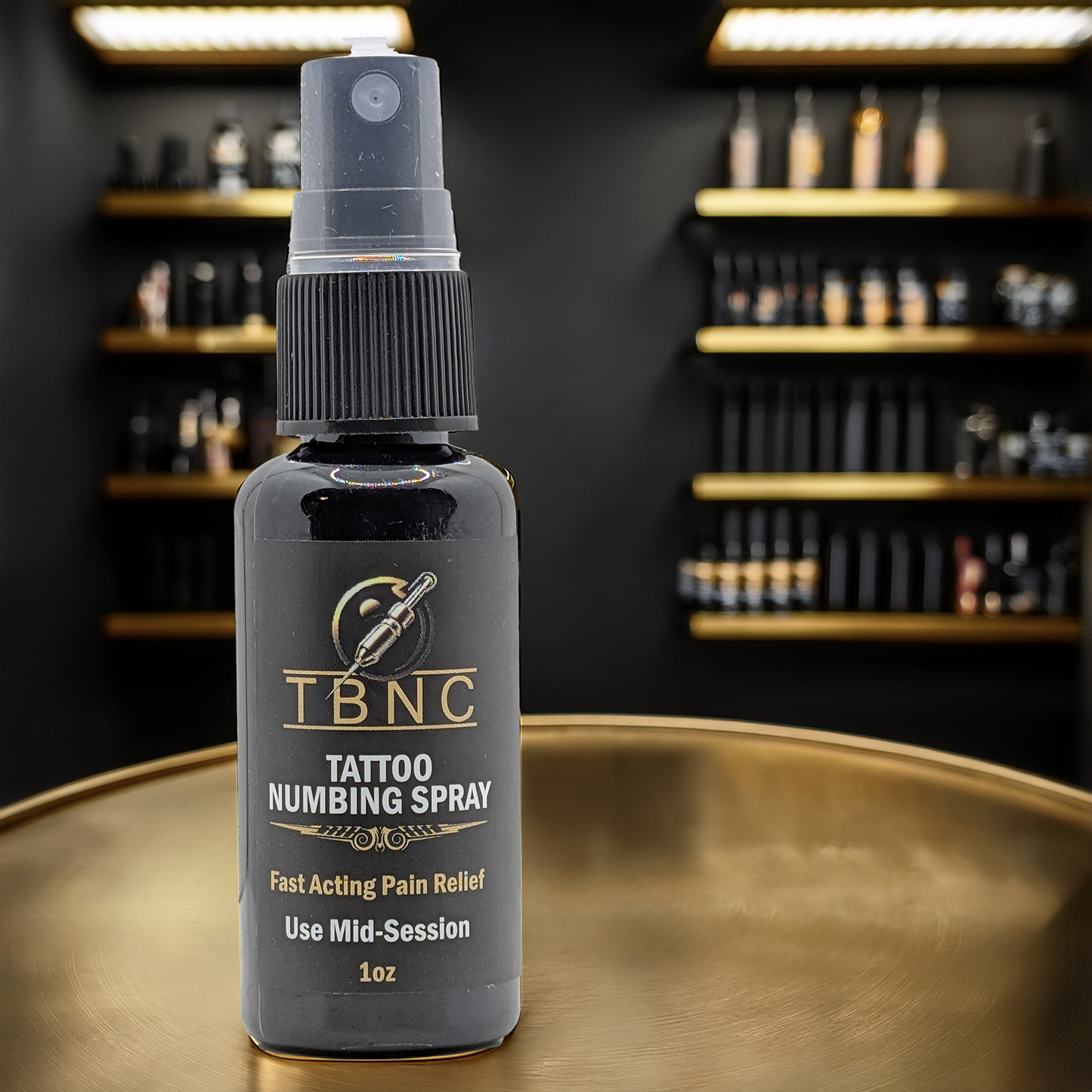 A bottle of TBNC - The Best Numbing Cream Tattoo Numbing Spray - Mid Session, known for fast-acting, maximum strength lidocaine pain relief, rests on a round gold surface with blurred tattoo ink bottles in the background.
