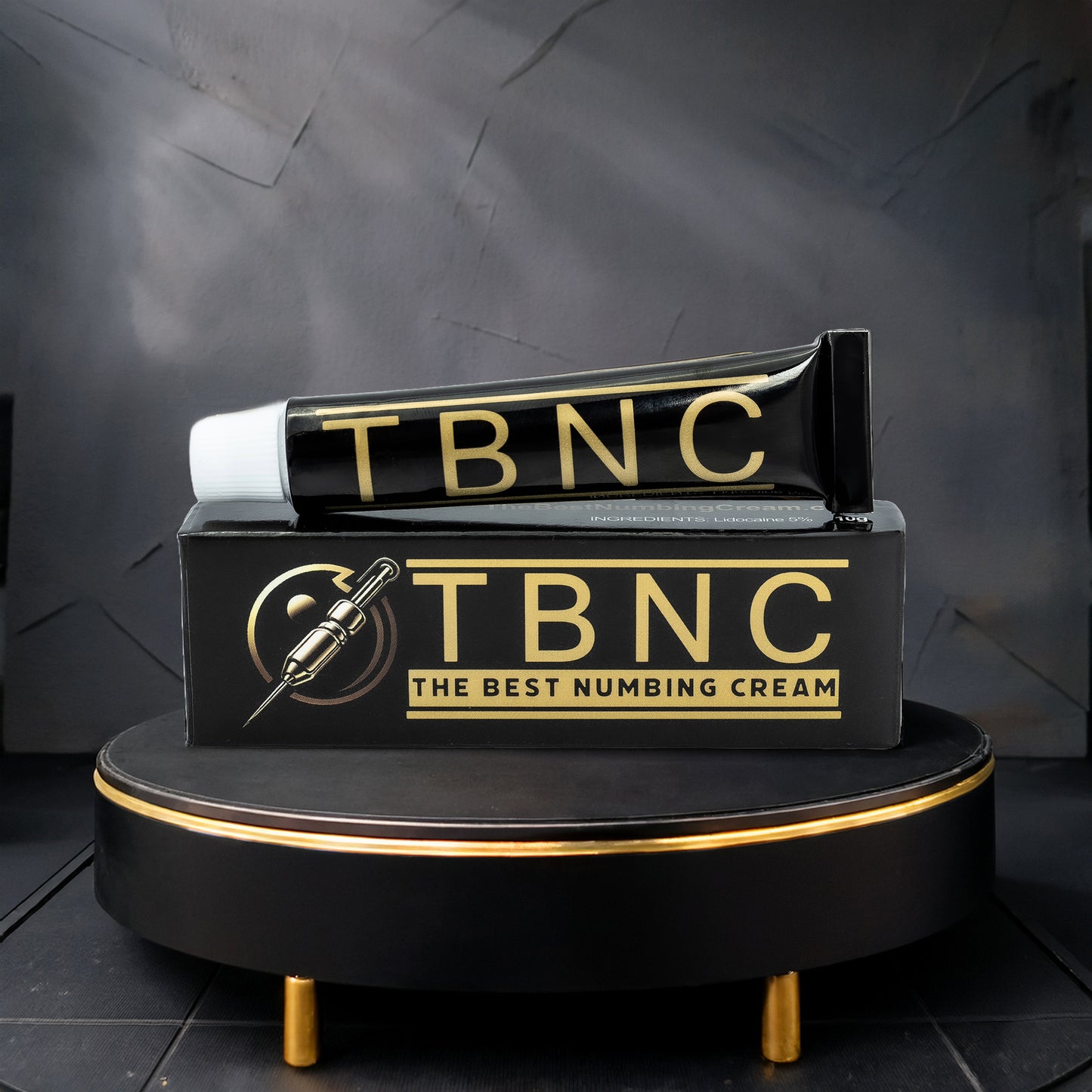 A black and gold "TBNC - The Best Numbing Cream" tube sits on its matching box with a needle icon, atop a round black platform—ideal for pain-free tattooing with this top Tattoo Numbing Cream.
