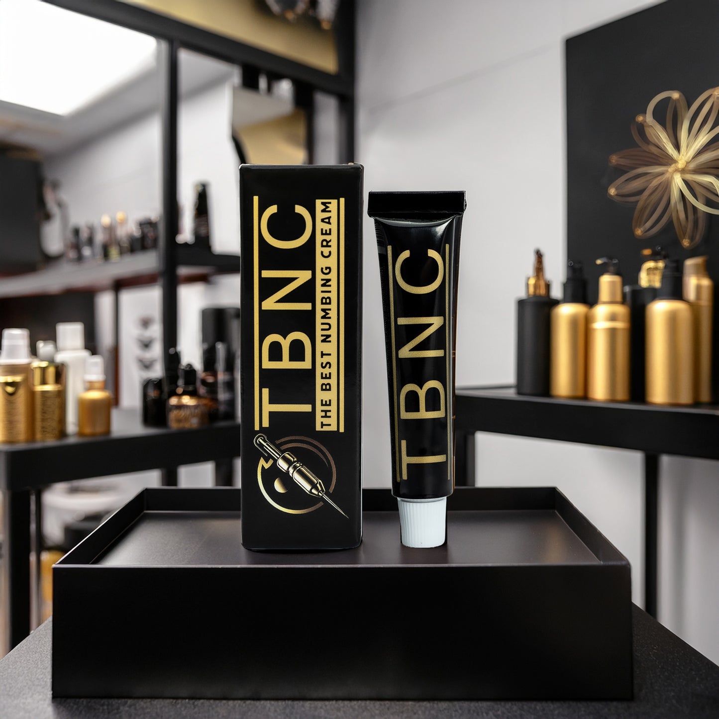A tube and box of Tattoo Numbing Cream by TBNC - The Best Numbing Cream, with black and gold packaging, sit on a black tray. Behind them, shelves display matching bottles, emphasizing pain-free tattoo sessions.