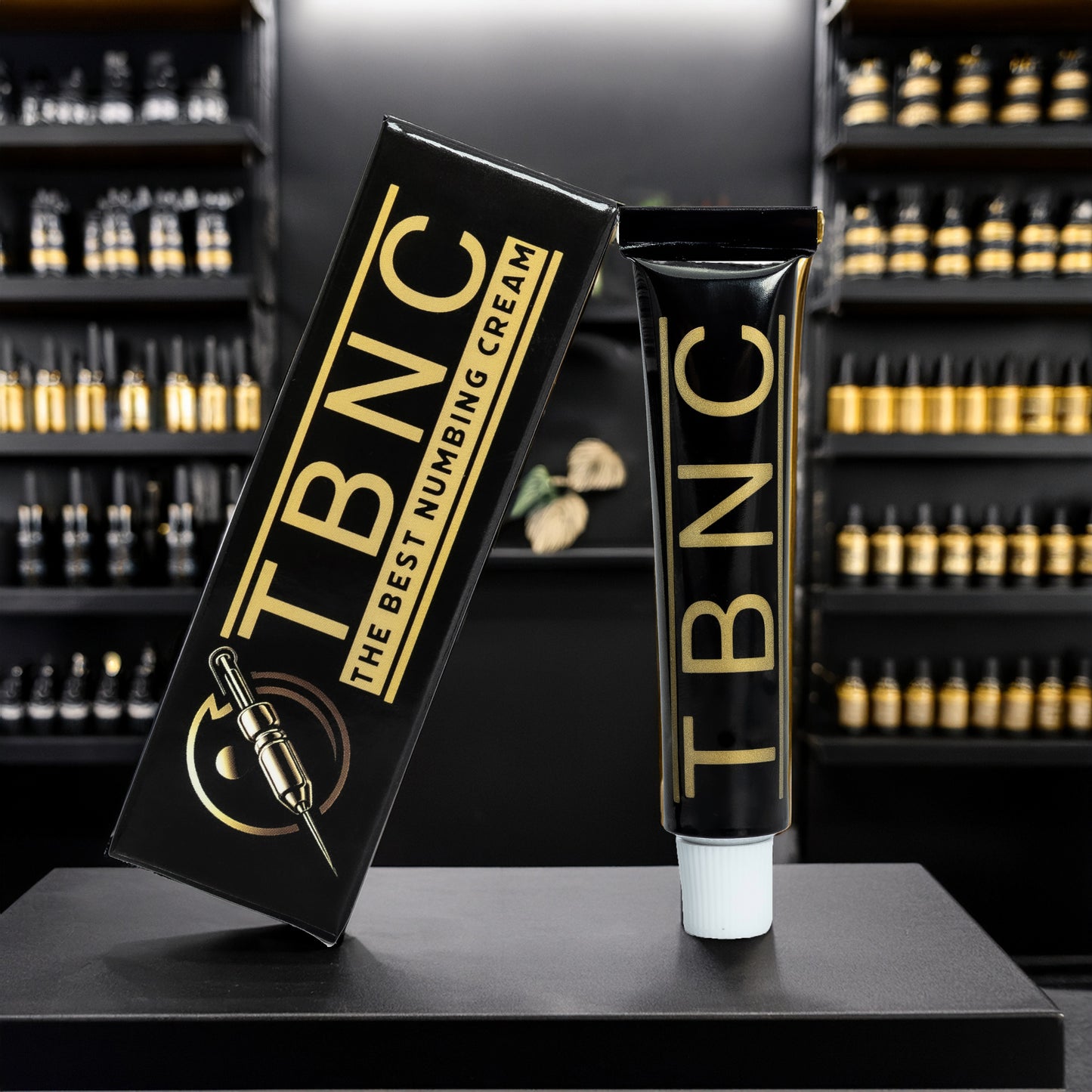 A black and gold tube labeled "TBNC" stands upright beside its box reading "The Best Numbing Cream," displayed on a dark surface with blurred shelves behind—ideal for pain-free tattooing with TBNC Tattoo Numbing Cream.