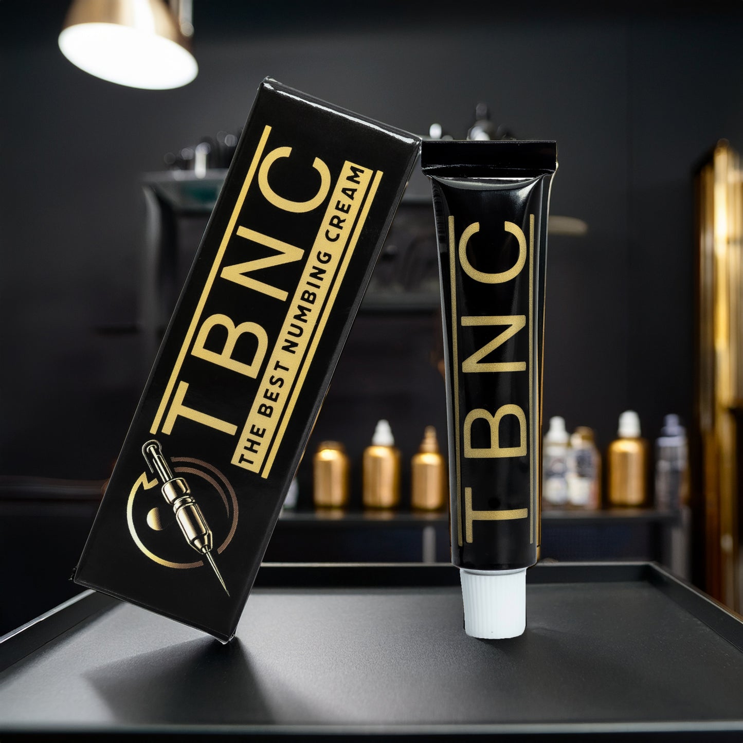 A sleek black and gold tube of TBNC - The Best Numbing Cream - 10 Pack sits by its box, branded with "The Best Numbing Cream" and a tattoo machine graphic—ideal for pain-free tattoos in a modern, dark-toned studio.