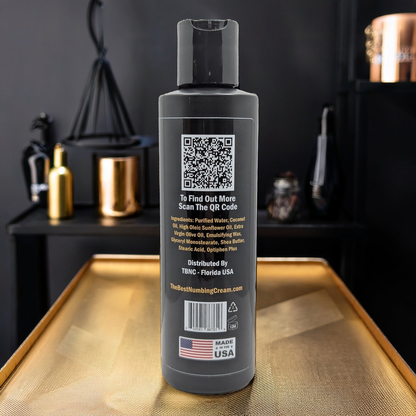 A black bottle of TBNC - The Best Numbing Cream Tattoo Aftercare Lotion - Unscented with a flip-top lid stands on a reflective gold surface. The label features ingredients, a QR code, and "Made in USA". Gold and black decor is blurred in the background.