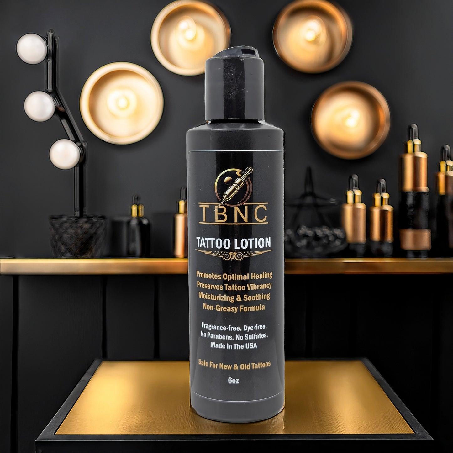 A black bottle of TBNC - The Best Numbing Cream Tattoo Aftercare Lotion - Unscented is shown on a reflective surface with gold and black decor, round lights, and tattoo ink bottles blurred in the background. The label highlights healing, moisturizing, and skin regeneration benefits.