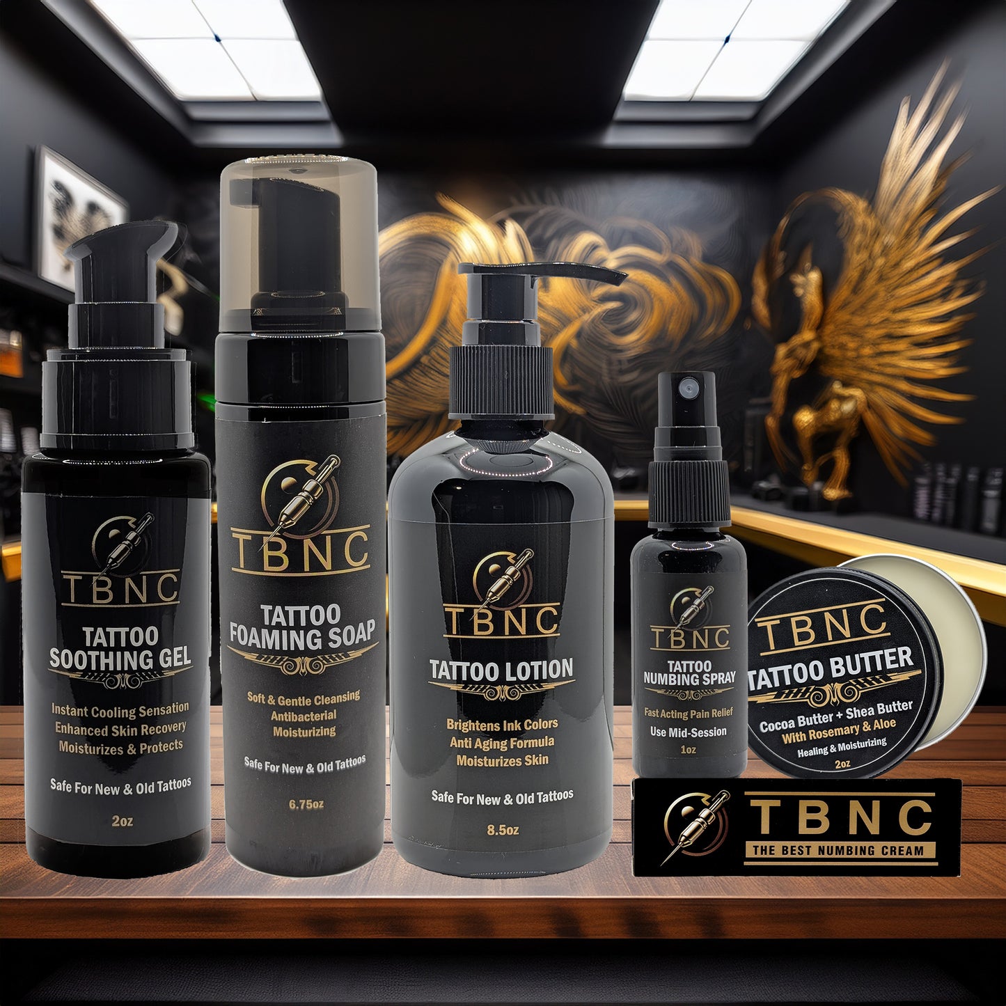 The TBNC - The Best Numbing Cream Tattoo Numbing & Aftercare Bundle, featuring five products, is displayed in a tattoo studio with black and gold décor and a large golden winged artwork on the wall behind.