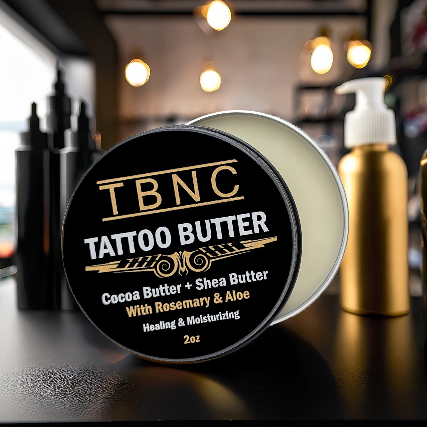 An open tin of TBNC - The Best Numbing Cream Tattoo Butter sits on a counter, infused with cocoa butter and aloe for tattoo aftercare and deep hydration, surrounded by blurred lights.