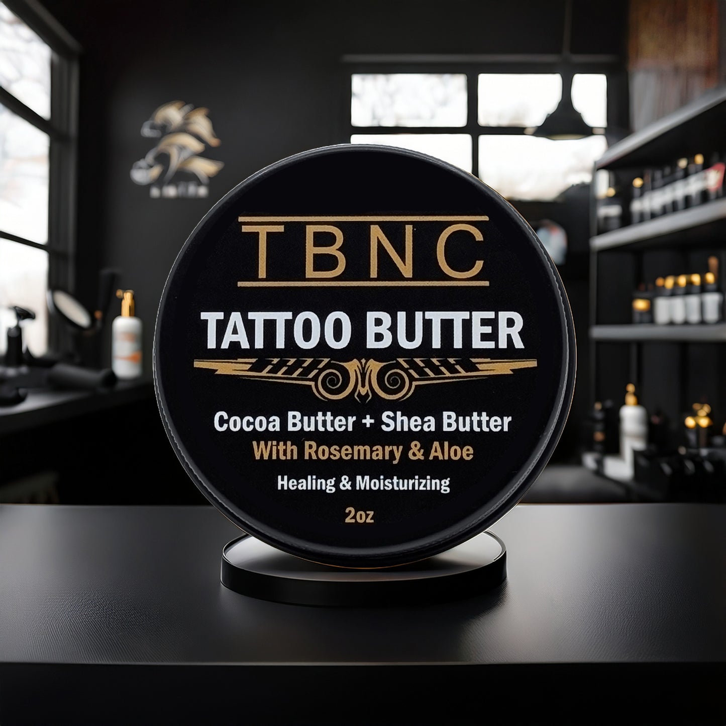A round container labeled “Tattoo Butter” by TBNC - The Best Numbing Cream sits on a stand in a modern tattoo studio, highlighting premium aftercare with cocoa butter, shea butter, rosemary, and aloe. Shelves and tattoo equipment are blurred in the background.