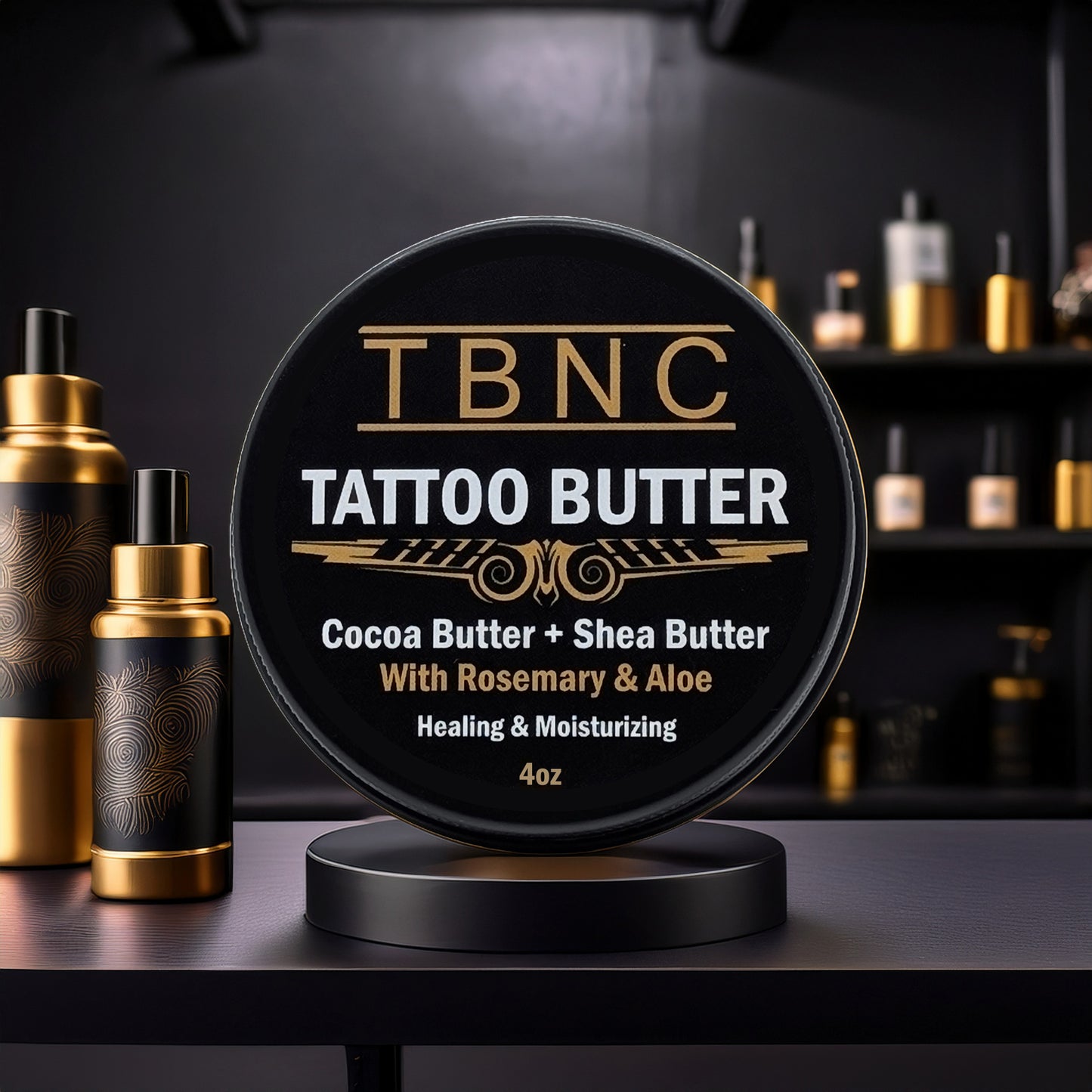 A black tin labeled "Tattoo Butter" by TBNC - The Best Numbing Cream sits on a stand. This 4oz blend with cocoa butter, shea butter, rosemary, and aloe soothes healing skin and locks in moisture for optimal tattoo aftercare. Gold and black bottles blur behind it.