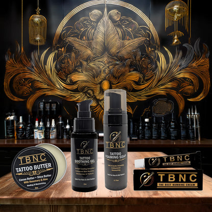 The TBNC - The Best Numbing Cream Beginner Tattoo Bundle, featuring numbing cream, soothing gel, foaming soap, and butter, is displayed in front of a gold and black ornate mural in a tattoo studio.