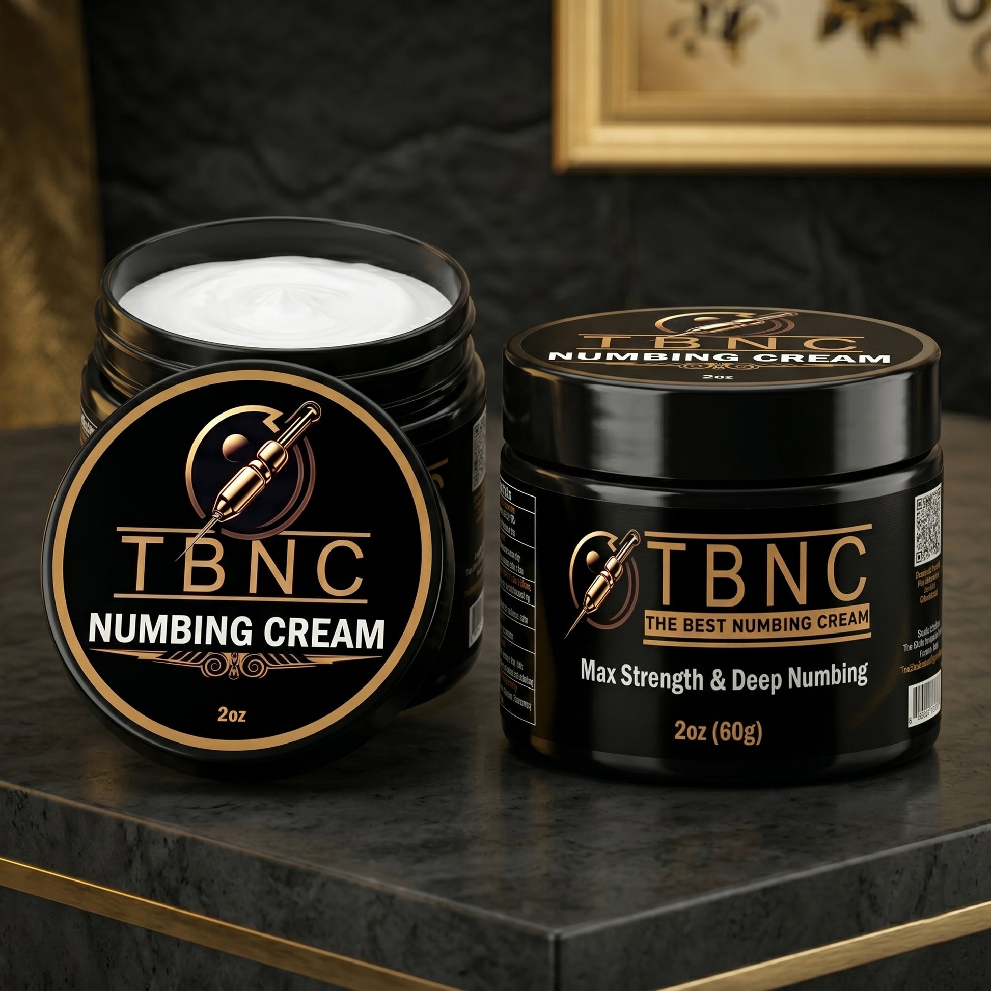 Two sleek black jars of bestselling TBNC Tattoo Numbing Cream MAX, one open to show the white cream, on a dark marble surface.