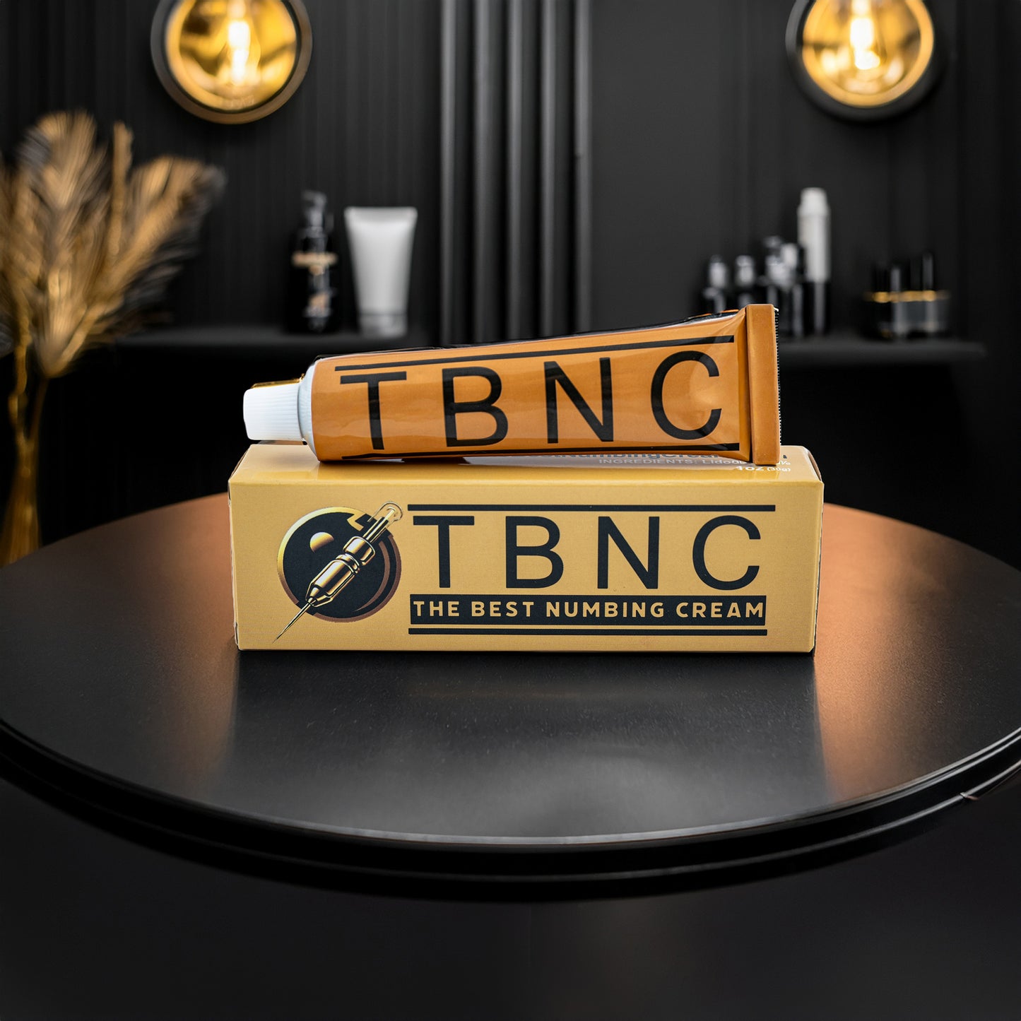 A tube and box labeled "TBNC - The Best Numbing Cream" Tattoo Numbing Cream XL - 2 Pack sit on a black table with modern decor and gold lighting in the background.