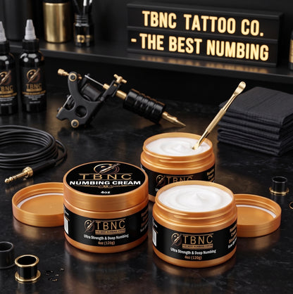 Premium Tattoo Numbing Cream ULTRA jars, gold spatula, pro tattoo gear, and towels on black counter. Branded max-strength sign.