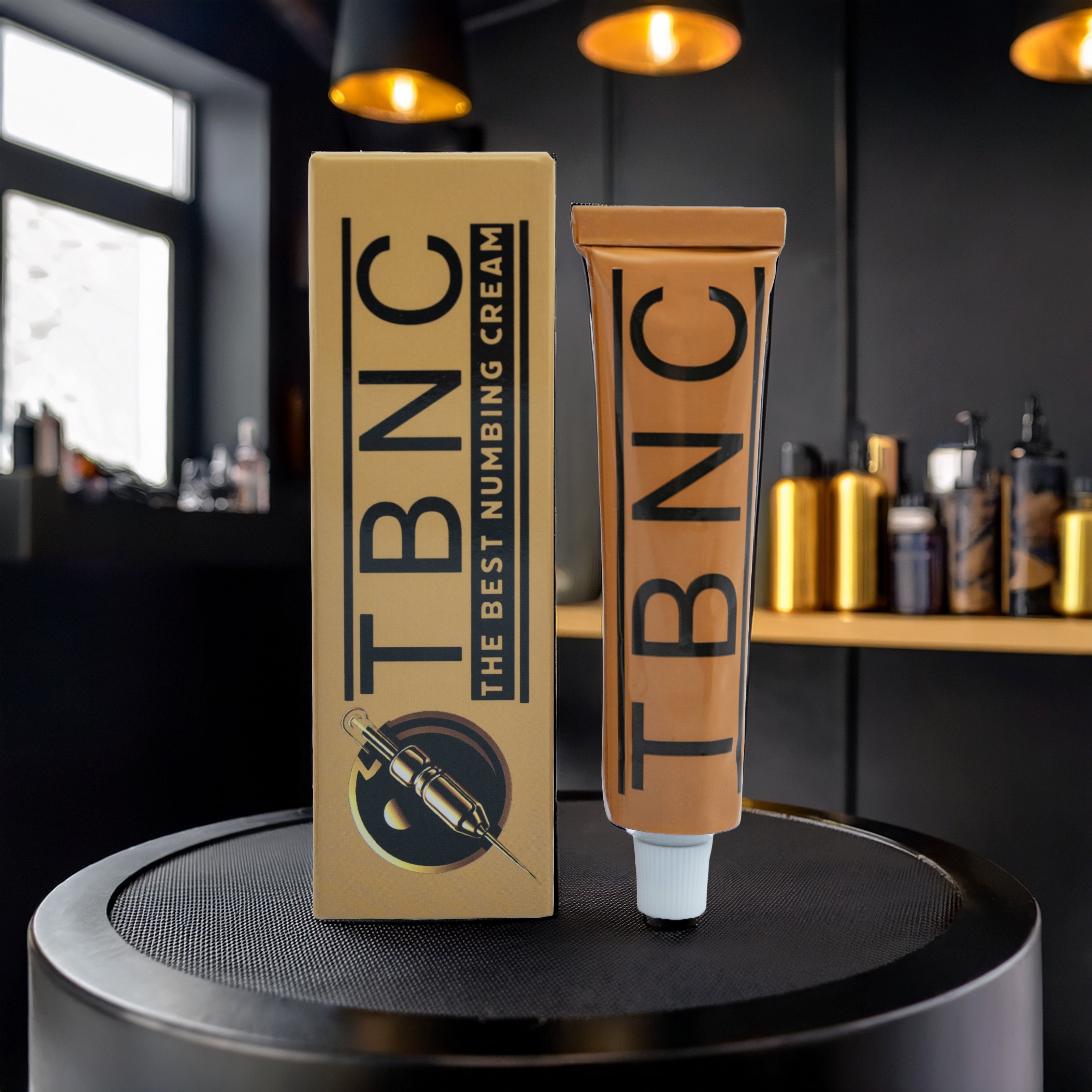 A tube and box of TBNC - The Best Numbing Cream's Tattoo Numbing Cream XL, a tattoo artist-approved numbing cream, sit on a black surface in a sleek, dark room with shelves of beauty products in the background.
