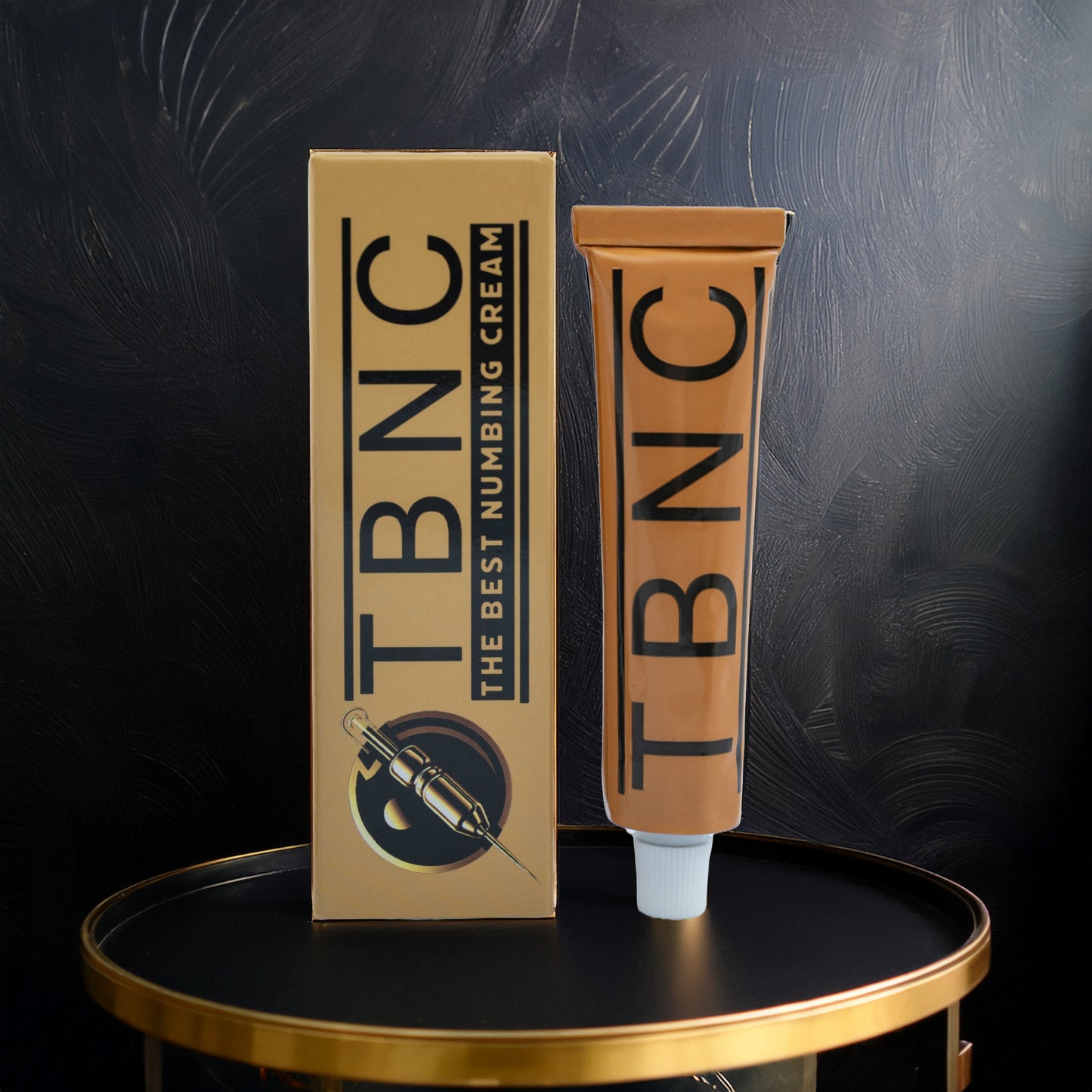 A tube and box of TBNC - The Best Numbing Cream Tattoo Numbing Cream XL with bold black lettering and a syringe illustration stand on a round black and gold table, the packaging highlighted against a dark textured background.