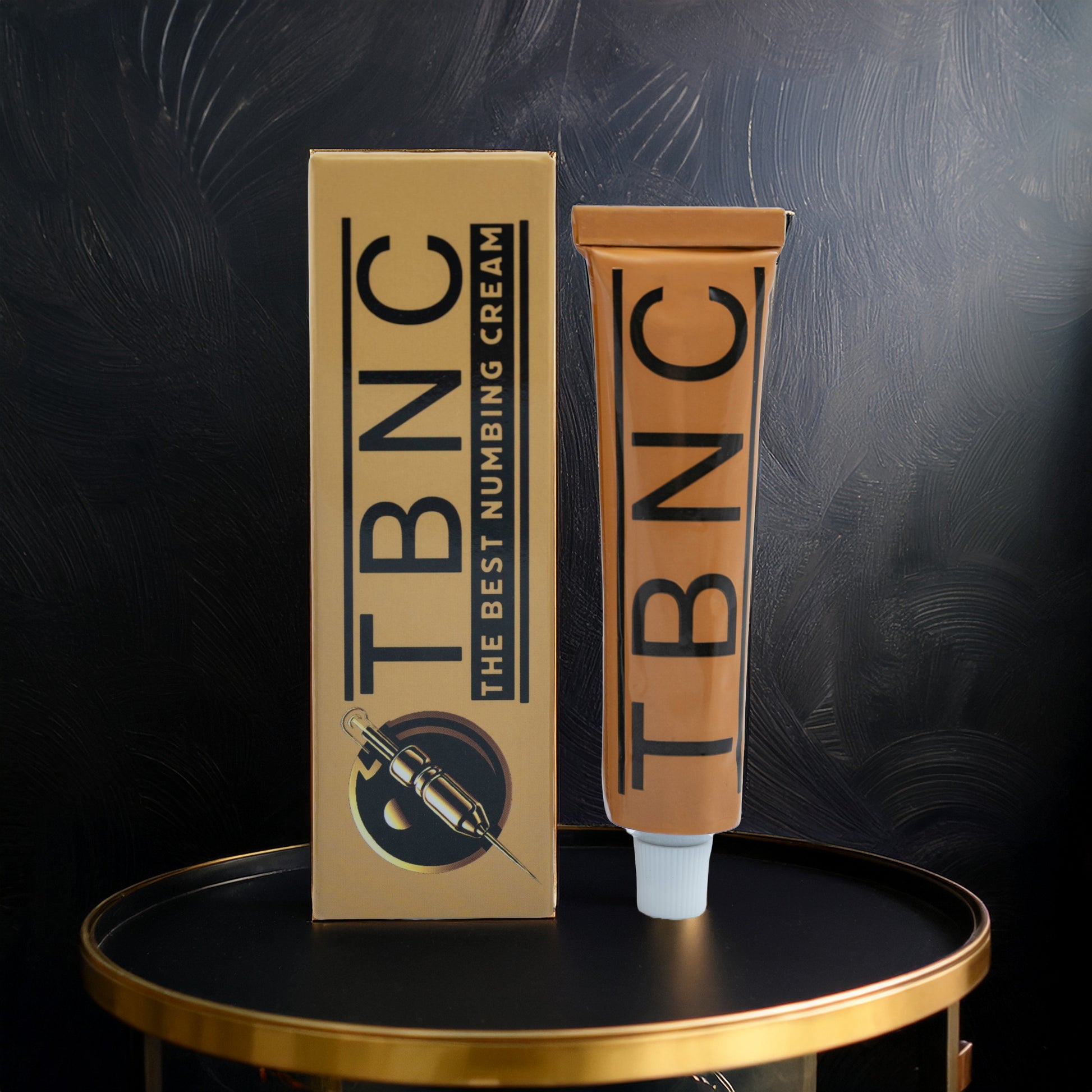 A tube and box of TBNC - The Best Numbing Cream Tattoo Numbing Cream XL with bold black lettering and a syringe illustration stand on a round black and gold table, the packaging highlighted against a dark textured background.