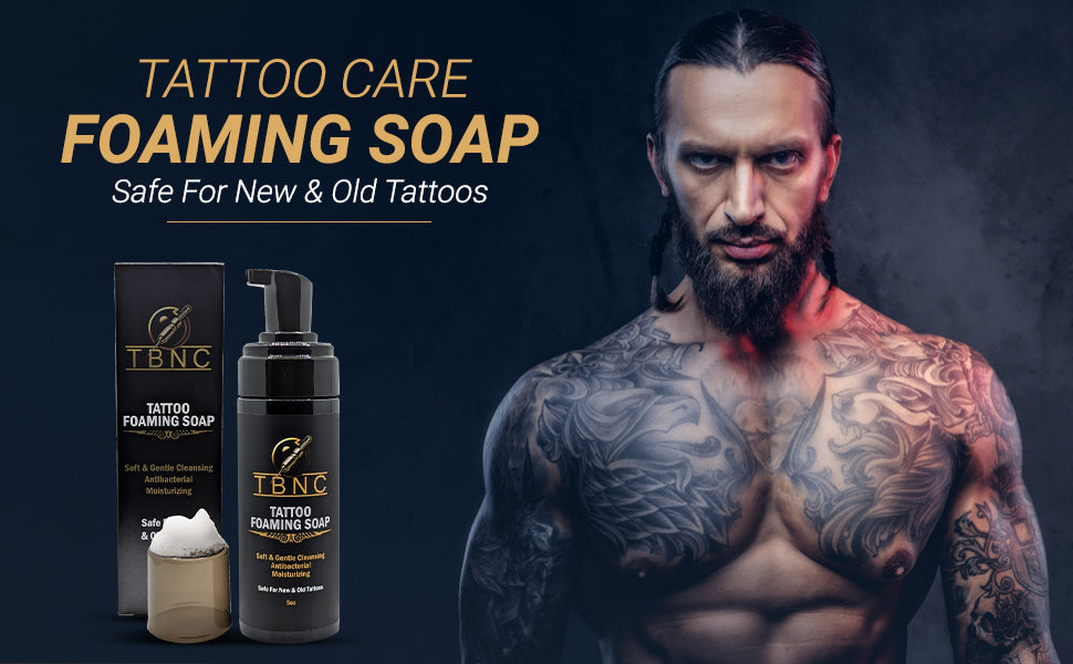 A muscular, tattooed man with long hair stands beside TBNC - The Best Numbing Cream Tattoo Foaming Soap bottles and packaging. Text reads: "Tattoo Foaming Soap—Antibacterial Care for New & Old Tattoos" on a dark background.
