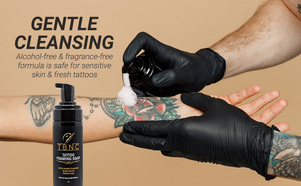 A person in black gloves dispenses TBNC - The Best Numbing Cream Tattoo Foaming Soap onto a tattooed arm, highlighting its gentle, alcohol-free, and fragrance-free formula. The product bottle is shown in the lower left corner.