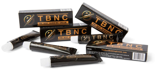 Several boxes and tubes of TBNC - The Best Numbing Cream - 4 Pack are arranged in a scattered style. The black packaging with gold text and a logo depicting a needle stands out, making it perfect for pain-free tattoos. The website "TheBestNumbingCream.com" is visible on one of the boxes.