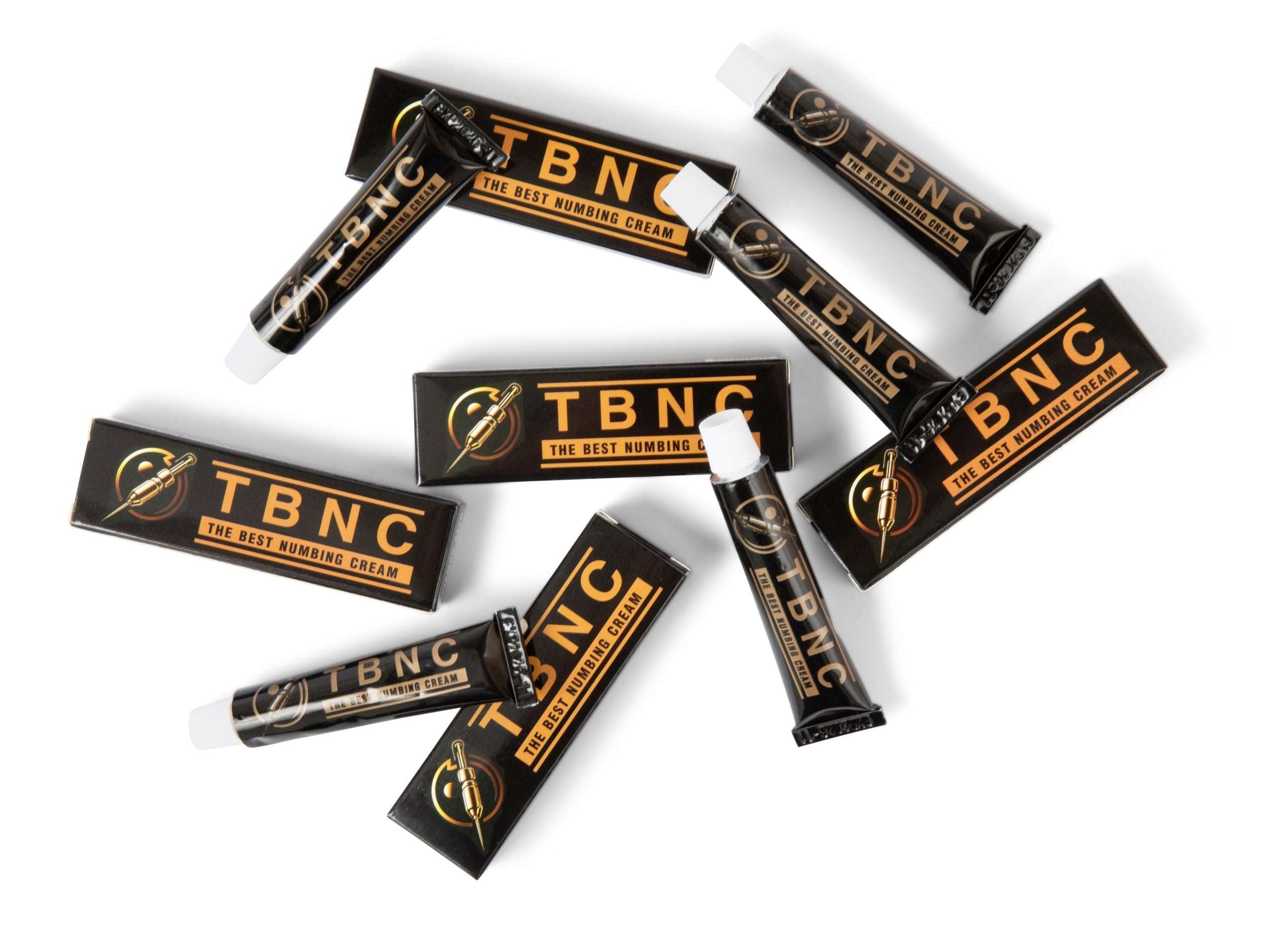 Image of several tubes and boxes of The Best Numbing Cream - TBNC in black and orange packaging scattered on a white background. The tubes, designed for painless tattoos, have white caps, and the text is prominently displayed on both the tubes and boxes.