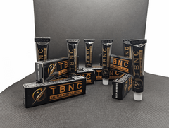 The image showcases multiple black tubes and boxes of "The Best Numbing Cream - TBNC - 6 Pack" from the brand TBNC. Arranged on a plain gray background, the tubes are labeled with "The Best Numbing Cream" and feature advanced numbing properties. Some tubes are standing while others are placed next to or atop their respective packaging boxes. Ideal for ensuring painless tattoos.