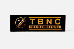 A rectangular product box with a black background showcases the TBNC logo and text in large gold letters, accompanied by "THE BEST NUMBING CREAM" in smaller gold text beneath it. An illustration of a needle inside a circle is positioned to the left of the text, emphasizing its advanced numbing properties for painless tattoos.