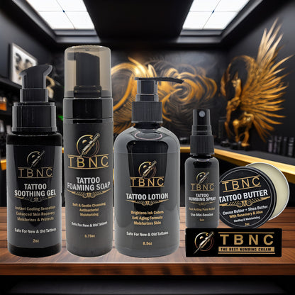 The TBNC - The Best Numbing Cream Tattoo Numbing & Aftercare Bundle, featuring five products, is displayed in a tattoo studio with black and gold décor and a large golden winged artwork on the wall behind.