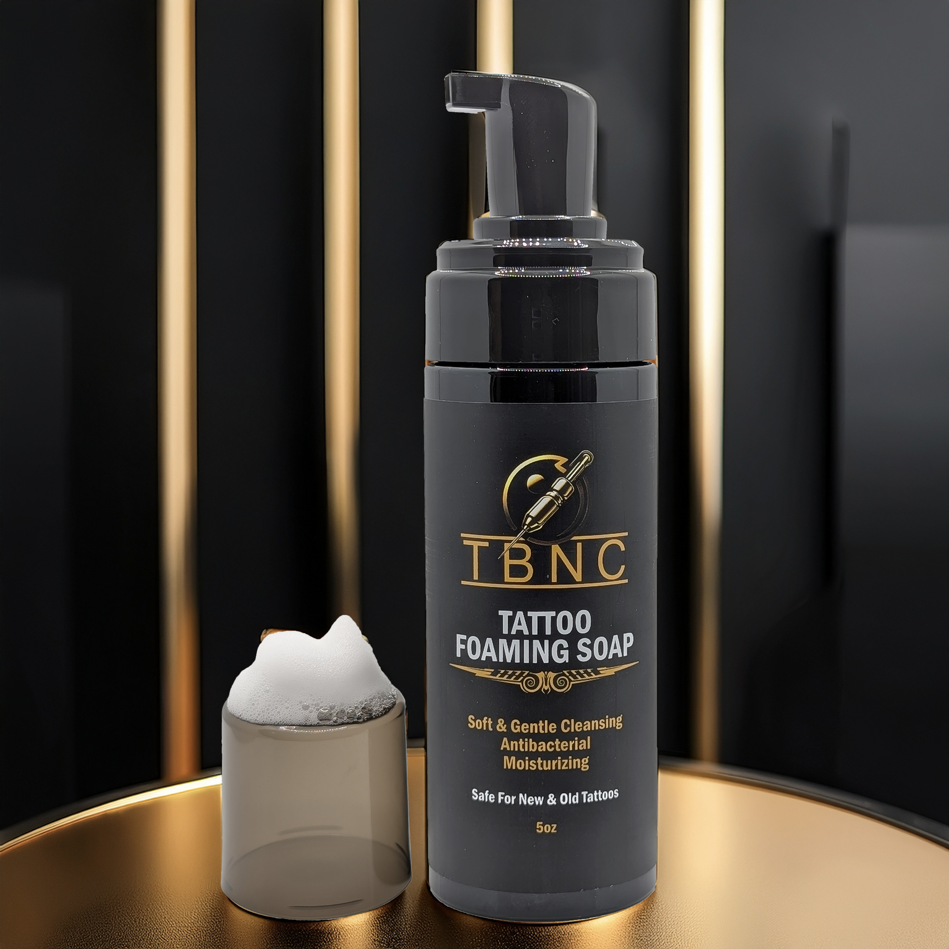 A bottle of TBNC - The Best Numbing Cream Tattoo Foaming Soap sits on a round surface next to a glass with foam. Its label highlights “Soft & Gentle Cleansing, Antibacterial, Moisturizing,” set against black with gold accents.