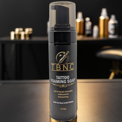 A close-up of a black TBNC Tattoo Foaming Soap bottle with a gold logo on a dark surface promises soothing, hydrating, and antibacterial benefits. A blurred background with shelves of similar bottles and a dimly lit chair enhance the ambiance.