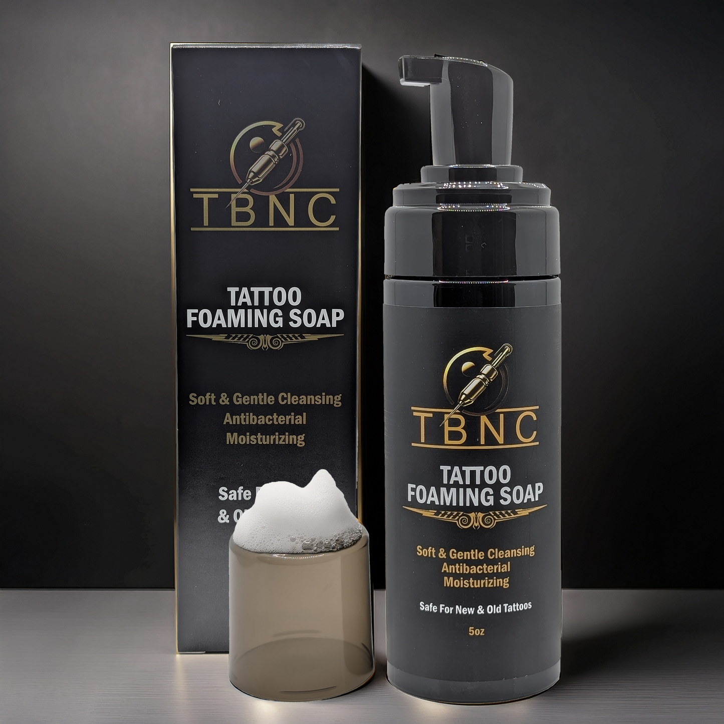 A bottle of TBNC - The Best Numbing Cream Tattoo Foaming Soap with a pump dispenser stands beside its black and gold box, with a small clear cup of foam in front. This antibacterial cleanser is ideal for tattoo aftercare.