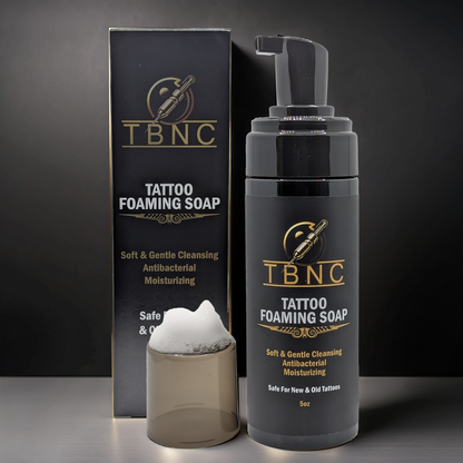 A bottle of TBNC - The Best Numbing Cream Tattoo Foaming Soap with a pump dispenser stands beside its black and gold box, with a small clear cup of foam in front. This antibacterial cleanser is ideal for tattoo aftercare.