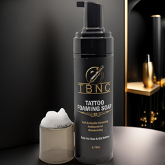 A TBNC - The Best Numbing Cream Tattoo Foaming Soap bottle with a pump top is shown beside a clear cap filled with foam. The label highlights antibacterial protection for both new and old tattoos, offering soothing and hydrating care against a sleek, modern backdrop.