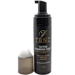 A black TBNC Tattoo Foaming Soap bottle with a gold logo, cap off and foam spilling out, showcases its gentle cleansing properties. It offers antibacterial protection and moisturizing benefits, making it safe for new or old tattoos and perfect for enhancing the healing process. Contains 6.75 oz.